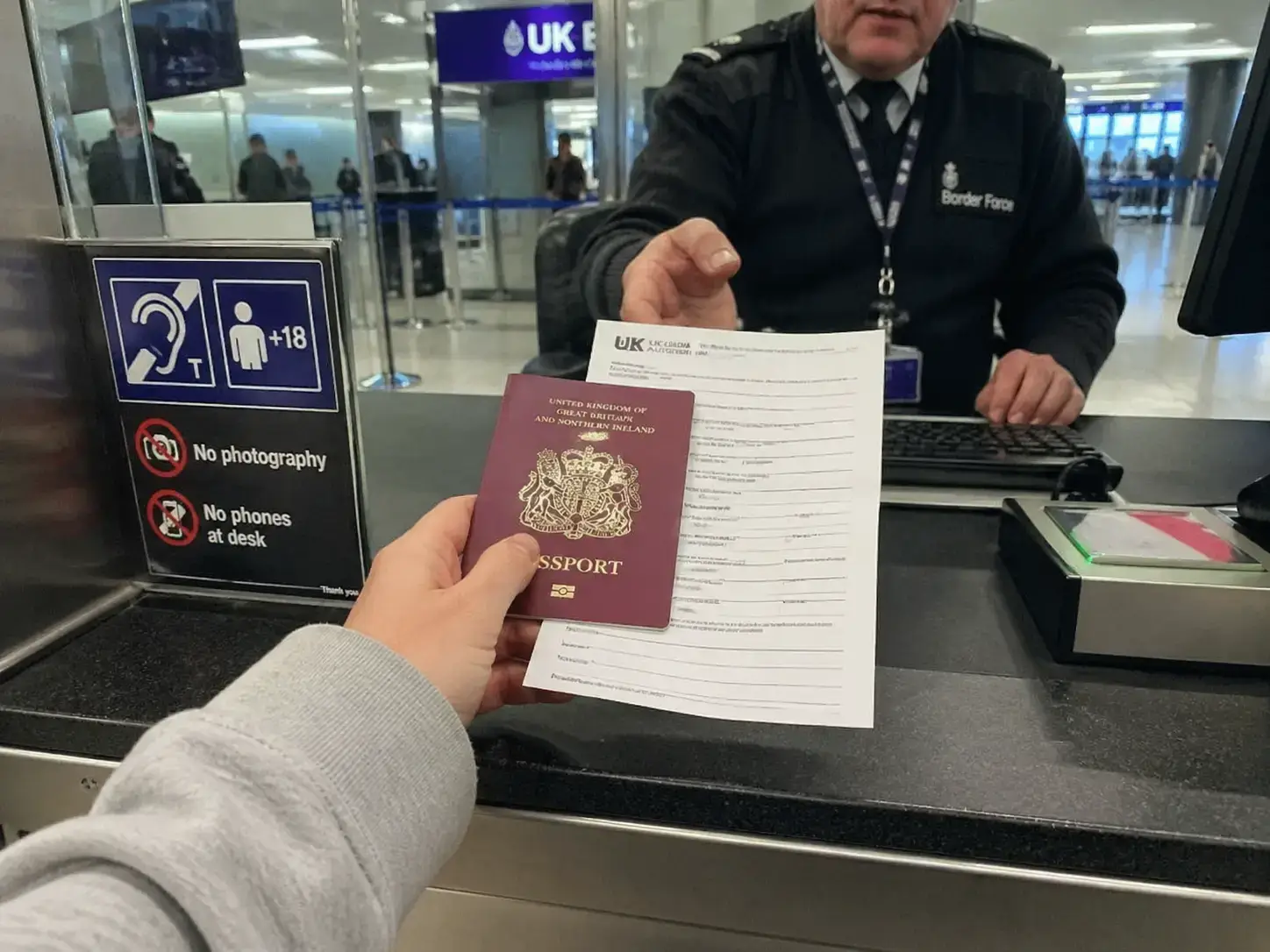 Uma fotografia documental vista por cima do ombro a mostrar um jovem viajante a entregar um passaporte e a autorização de viagem a um agente de controlo de fronteiras no aeroporto.
