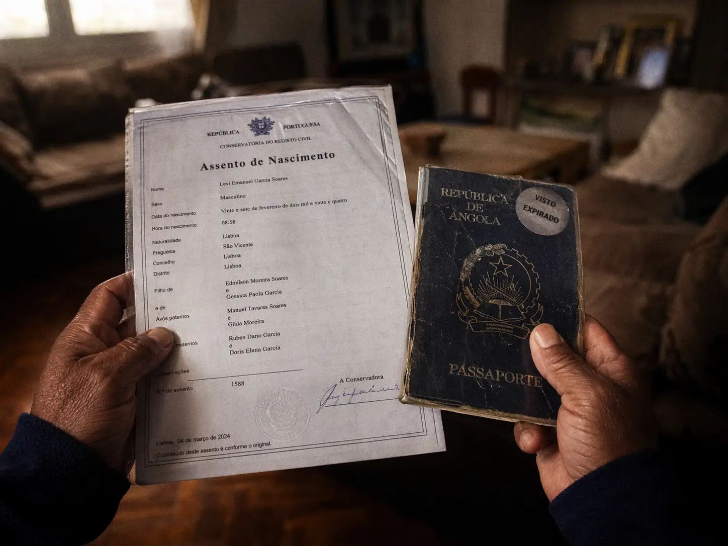Uma fotografia em grande plano de mãos a segurar uma certidão de nascimento portuguesa ao lado de um passaporte estrangeiro, ilustrando o contraste legal entre a criança e os pais.